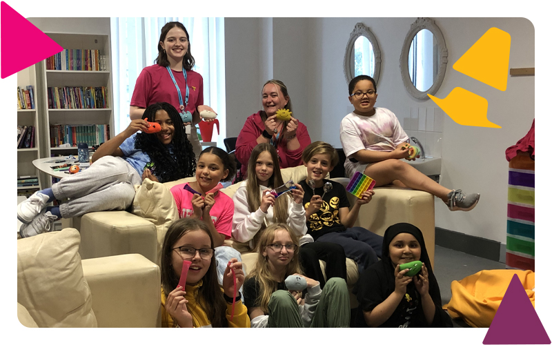 A group of young people and youth workers sitting on and around a sofa in the health and wellbeing room, holding colourful fidget toys