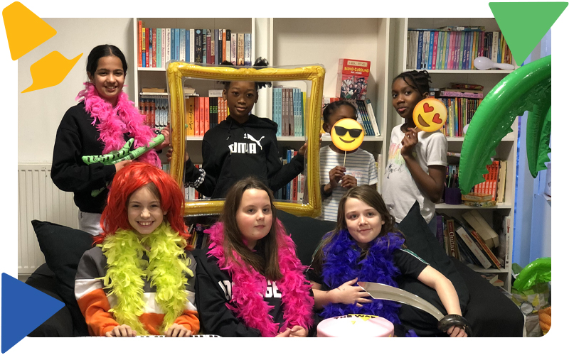 A group of young girls sitting on a black sofa wearing colourful feather boas and emoji faces