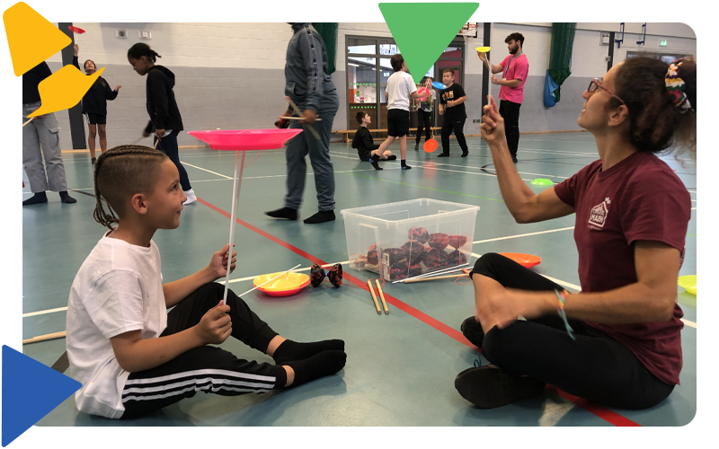 A young boy in a white t-shirt sitting on the floor of the sports hall with a lady in a red t-shirt who is teaching him to spin a red plate on a stick