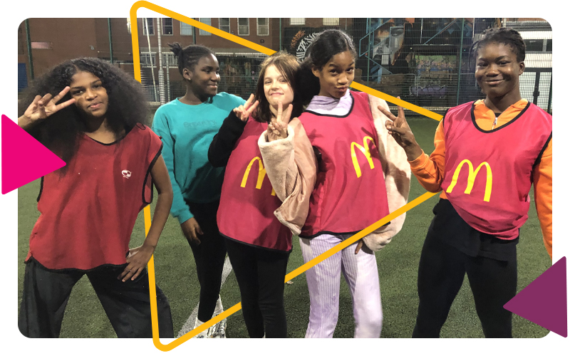 Five teenage girls wearing sports bibs on the MUGA and posing, making v signs